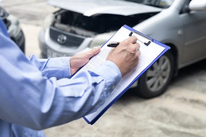 A man standing next to a wreck fills out information on a clipboard regarding car accident claims.