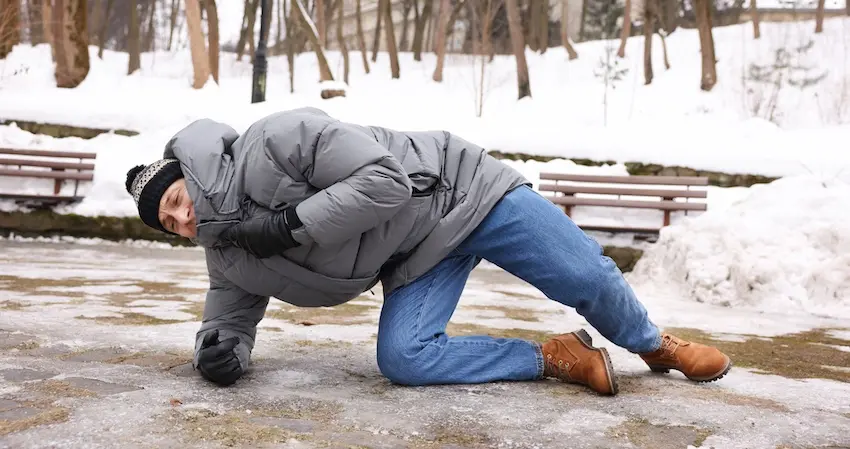 A person in a gray puffer jacket and winter hat struggles to get up after slipping and falling on an icy surface in a snow-covered park depicting the type of hazardous conditions that frequently lead to personal injury lawsuits against negligent property owners in South Carolina.