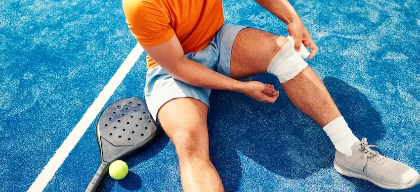 A male athlete in an orange shirt sits on a blue sports court holding his bandaged knee beside a paddle racket and tennis ball representing the kind of sports facility injury that may give rise to personal injury lawsuits when unsafe court conditions are a contributing factor in South Carolina.