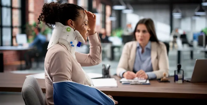 A woman wearing a neck brace and arm sling holds her hand to her forehead while seated across from a female attorney with a laptop in a modern office depicting an injured client discussing the details of her personal injury lawsuit during a legal consultation at McDougall LawFirm LLC in South Carolina.