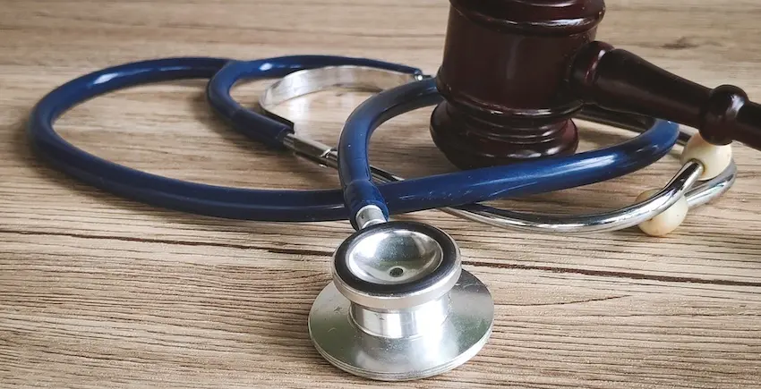 A blue stethoscope with a silver chest piece and white earbuds is coiled across a weathered wooden surface with a dark mahogany judge's gavel resting partially inside the loop of the tubing- both objects overlapping on the natural wood grain tabletop — a visual representation of the intersection between medical evidence and legal proceedings at the heart of every personal injury case with help from McDougall LawFirm LLC.