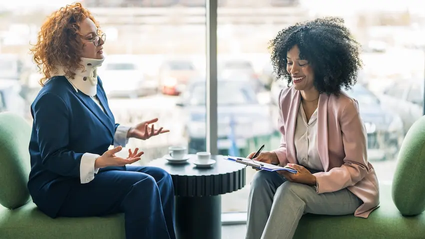 A woman with curly red hair wearing a navy blue blazer and a white cervical neck brace sits across from a professionally dressed attorney with voluminous curly hair in a pink blazer who is smiling and writing on a clipboard- the two seated on rounded green cushioned chairs at a small round black table with two white coffee cups in front of a large floor-to-ceiling window overlooking a sunlit parking lot — depicting the initial consultation and case evaluation stage with a lawyer from McDougall LawFirm LLC- of a personal injury case in South Carolina.