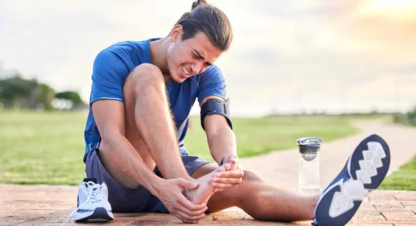 A young male runner with dark hair pulled back in a bun sits on a brick pathway gripping his ankle and lower leg with both hands- his face grimacing in visible pain- wearing a blue athletic shirt- dark shorts- white sneakers- and a black phone armband- with a clear water bottle beside him and a wide open green field and soft glowing sky stretching out behind him — the type of serious physical injury suffered due to another party's negligence that forms the basis of a personal injury case with help from McDougall LawFirm LLC in South Carolina.