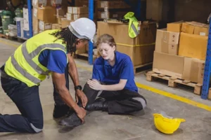 A man in a warehouse helping a woman who is suffering from knee pain after a work related injury and the woman is considering help from a personal injury lawyer.